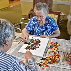 an older woman sitting at a table working on a tree art project with other people