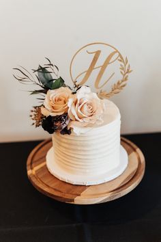 a white wedding cake with flowers and the letter b on top is sitting on a wooden platter