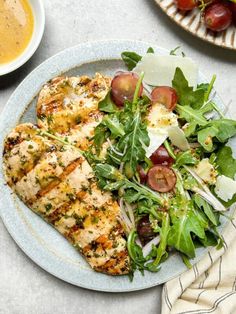 a white plate topped with chicken and salad next to a bowl of dressing on top of a table