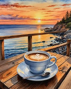 a cup of coffee sitting on top of a wooden table next to the ocean at sunset