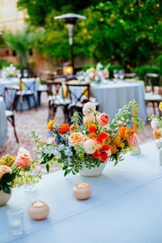 an arrangement of colorful flowers in vases on a white table cloth covered outdoor dining area