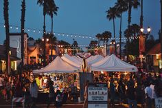 a crowd of people walking around a street next to tall palm trees and lights strung over the buildings