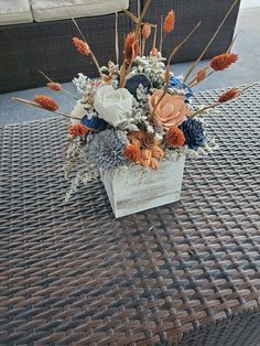 an arrangement of flowers in a square vase on a wicker patio table with outdoor furniture