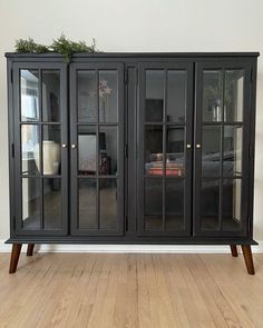 a black china cabinet with glass doors and wooden legs on the floor in front of a white wall