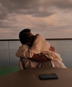 a person sitting at a table with their arms around each other and the ocean in the background