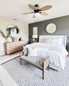 a white bed sitting in a bedroom next to a dresser and mirror with a fan on it