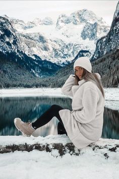 a woman sitting on the edge of a lake with snow covered mountains in the background