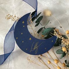 a paper moon and some dried flowers on a white surface with blue ribbon around it