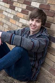 a young man sitting on the ground next to a brick wall with his hands in his pockets