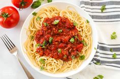 a white bowl filled with spaghetti and sauce next to two tomatoes on a tablecloth