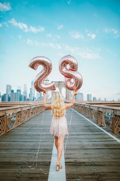 a woman walking across a bridge holding up two large balloons in the shape of numbers