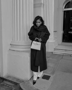 black and white photograph of woman in coat leaning against column with shopping bag on sidewalk