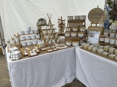 an assortment of products on display for sale at a farmers market table with white linen covered tables