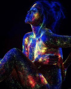 a woman is sitting in the dark with colored powder on her body and hands behind her back