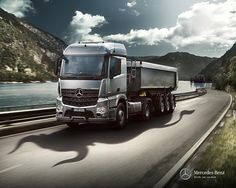 a large truck driving down a road next to a body of water with mountains in the background