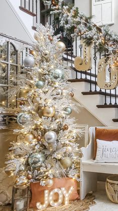 a white christmas tree with gold and silver ornaments in front of a stair case that says joy