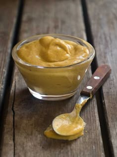 a small glass bowl filled with mustard on top of a wooden table next to a spoon