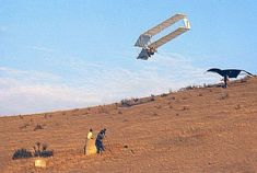 two people standing on top of a dry grass covered hill next to a flying glider