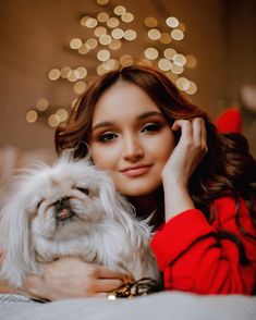 a woman holding a small white dog in her arms and looking at the camera with christmas lights behind her