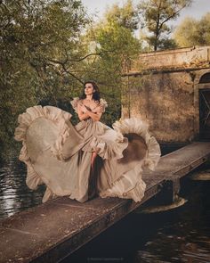 a woman in a long dress is sitting on a pier near the water and trees