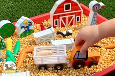 a child's hand is playing with toys in a red tray filled with corn