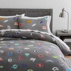 a bed with a gray comforter covered in lots of different sports logos on it