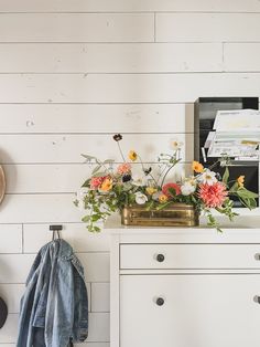 a white dresser topped with flowers next to a coat rack and wall mounted clock on top of it