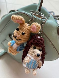 two crocheted keychains are hanging from a purse handle, one is holding a stuffed animal and the other is wearing a sweater