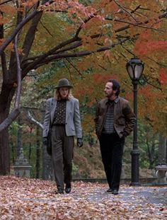 two people are walking in the park with leaves on the ground and one is wearing a hat