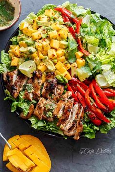 a large salad with chicken, mangoes and lettuce on it next to a bowl of dressing