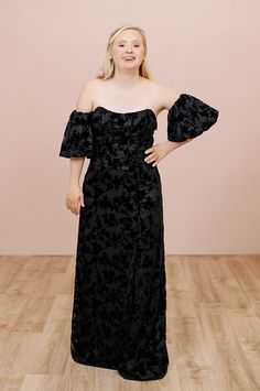 a woman standing on a wooden floor wearing a black dress with off the shoulder sleeves