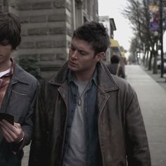 two young men are walking down the street looking at their cell phones and one is wearing a brown leather jacket