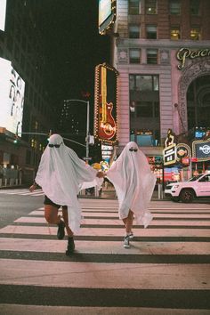 two women dressed in ghost costumes cross the street at an intersection with neon signs and buildings behind them