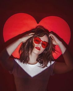 a woman with red heart shaped sunglasses on her head and hands behind her head, in front of a dark background