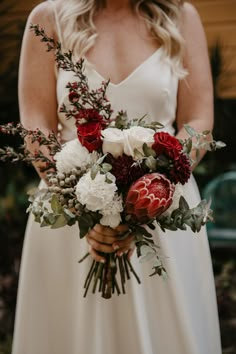 a woman in a white dress holding a bouquet of red and white flowers with greenery