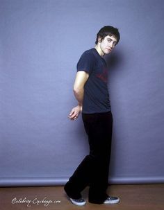 a young man standing in front of a wall with his hand on his hip and looking at the camera