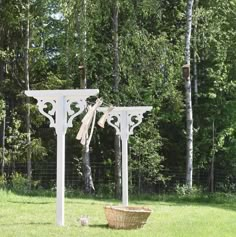 a white arbor in the middle of a grassy area with a basket under it and trees behind it