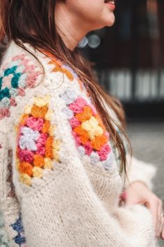 a woman wearing a white sweater with colorful crocheted flowers on it's sleeves