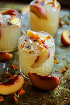 two glasses filled with ice and fruit on top of a metal table next to peaches