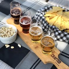 three glasses of beer are sitting on a wooden tray next to chips and tortillas