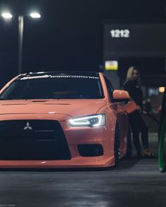 an orange car parked in front of a gas station at night with people standing around