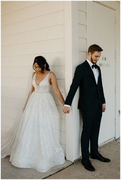 a bride and groom hold hands as they stand in front of a white wall holding hands