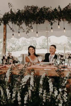 two people sitting at a table with flowers and greenery on the tables in front of them