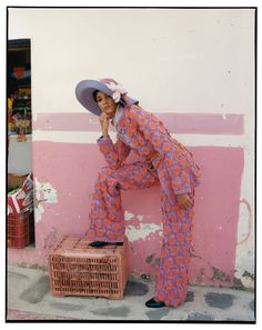 a woman leaning against a pink wall with a hat on her head and a wicker basket in front of her