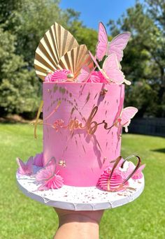 a pink and gold birthday cake with butterflies on it's top is being held by a person