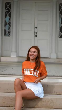 a woman in an orange shirt and white skirt sitting on some steps smiling at the camera