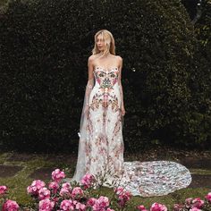 a woman standing in front of flowers wearing a white dress with floral designs on it