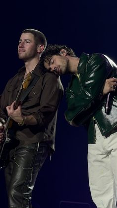 two men standing next to each other on stage with one holding a guitar and the other wearing a leather jacket