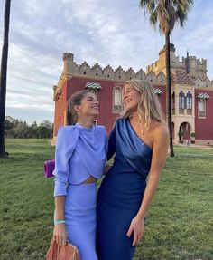 two women standing next to each other in front of a building with palm trees and grass