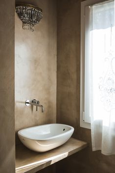 a bathroom sink sitting under a window in front of a white curtain and a chandelier